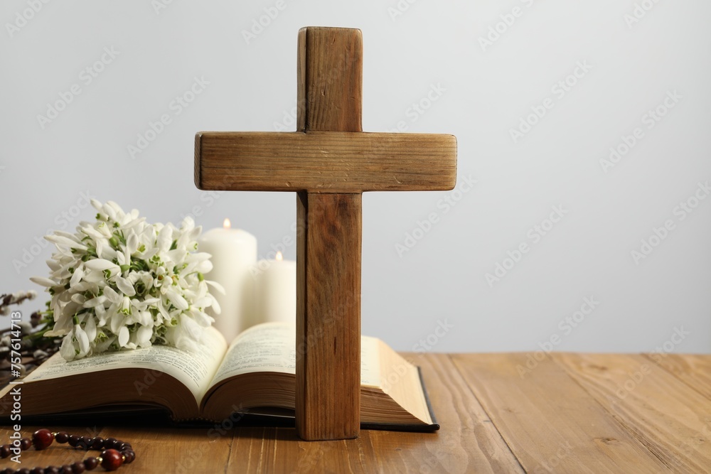 Cross, Bible, rosary beads, flowers and church candles on wooden table against light background. Space for text