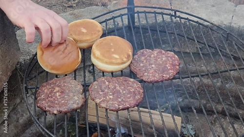 Close up of three deer burgers being grilled next to roasting buns over deep dire pit burning wood for deep rich smoke flavor. photo