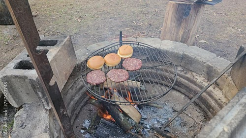 Three freshly made burgers being grilled next to roasting buns over deep fire pit burning wood for smoky flavor. photo