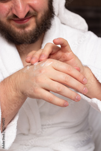Vertical photo. Young bearded man in white bathrobe applies cosmetic moisturizer to his male hands closeup in hotel room. Skin protection during cold season. Cosmetic procedure in spa salon. Hand care