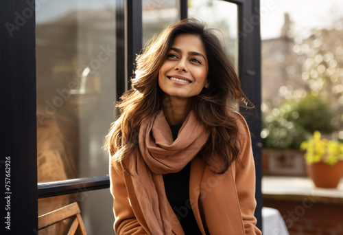 A 30s Indian woman scarf around her neck with long hair smiling sitting on the balcony outside a large glass door of a modern apartment brown coat over looking at the camera on a sunny day 