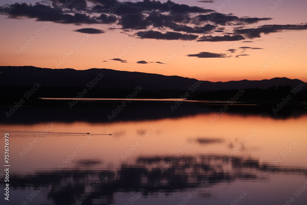 Colourful sunset over a flat lake with mountains in the background and clouds reflecting in water while birds swim