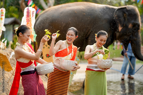 Closeup beautiful Thai young lady ware Thai traditional dress use flowers to sprinkle water on each other on the Thai New Year's Day in a fun way on blurred elephant and pile of sand background.
