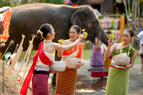Closeup beautiful Thai young lady ware Thai traditional dress use flowers to sprinkle water on each other on the Thai New Year's Day in a fun way on blurred elephant and pile of sand background.