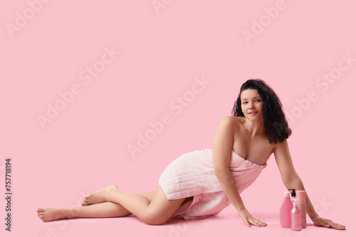 Young woman in towel after shower with cosmetic products lying on pink background