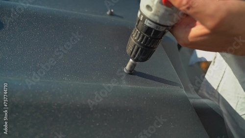 A roofer screws a self-tapping screw into a black metal tile using a screwdriver. Screwing a self-tapping screw into a metal roof close-up. Roof construction