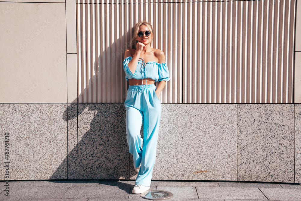 Young beautiful smiling hipster woman in trendy summer clothes. Sexy carefree woman posing in the street at sunset. Positive blond model outdoors. Cheerful and happy. Near wall. In sunglasses Young beautiful smiling hipster woman in trendy summer clothes. Sexy carefree woman posing in the street at sunset. Positive blond model outdoors. Cheerful and happy. Near wall. In sunglasses