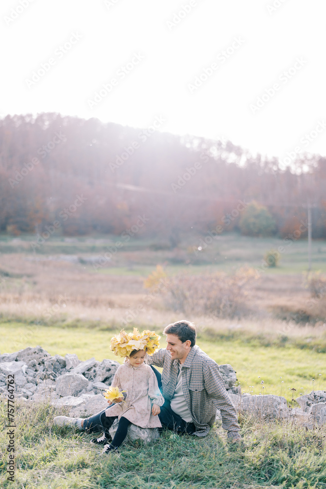 Little girl in a wreath of yellow leaves sits on a stone next to her dad on the sunny lawn