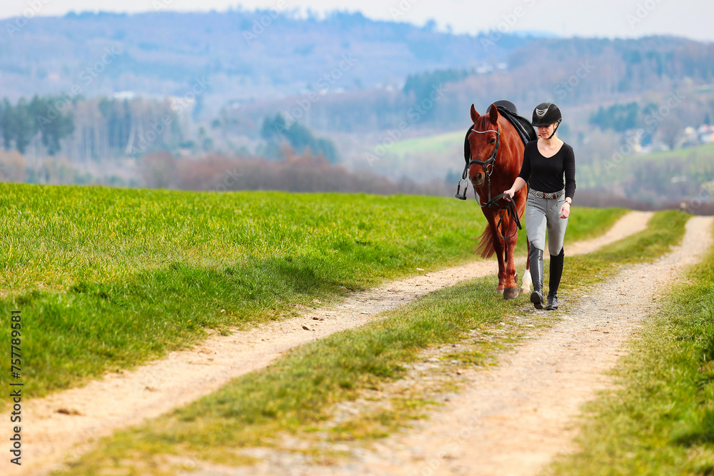 Obraz premium Horse and rider, saddled in riding clothes, walk side by side up a dirt road through green fields.