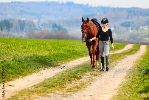 Horse and rider, saddled in riding clothes, walk side by side up a dirt road through green fields. © RD-Fotografie