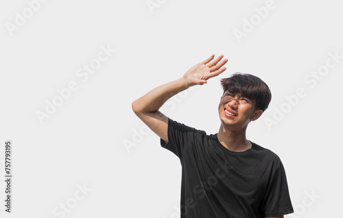 A young Asian man in his 20s wearing a black t-shirt is using his hands to block the sunlight that hits him, isolated on a gray background. The concept of facing the sun or hot weather