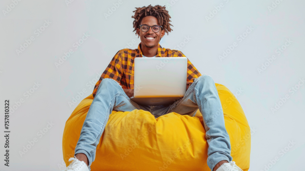 A joyful person sits cross-legged on a yellow beanbag, working on a laptop with a smile. - obrazy, fototapety, plakaty 