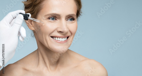 Cosmetologist using pipette dropper applies essential oil to face of mature woman for hydrated skin. Facial skin care with treatment essential oil and serum