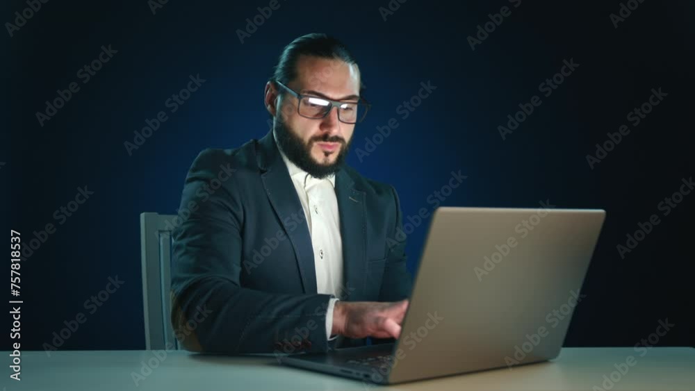 Confident bearded businessman in a black suit working on a laptop, focused expression, isolated on dark blue backdrop. Ideal for corporate digital communication themes. Camera 8K RAW. 
