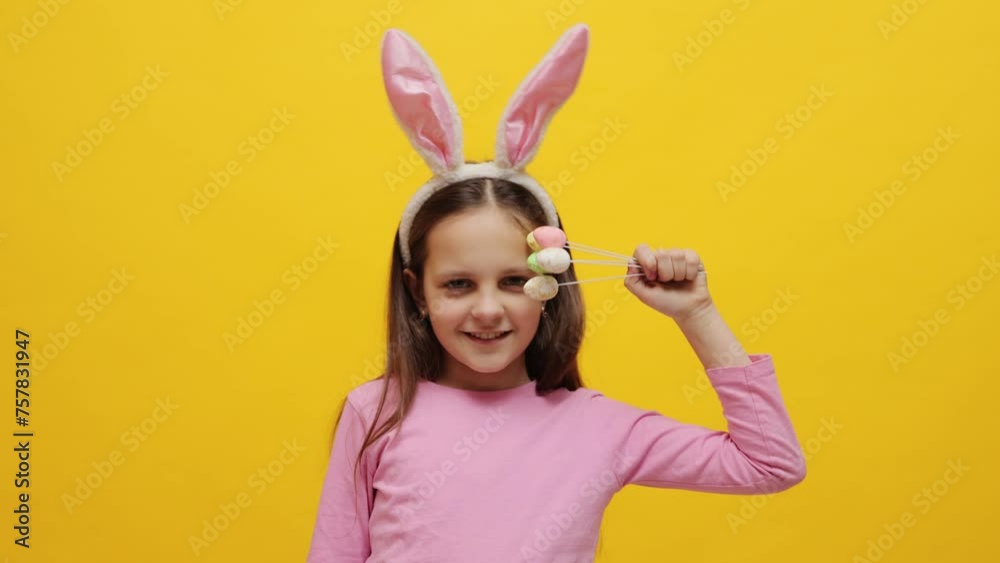 Extremely happy little girl wearing pink bunny ears headband holding pop cakes standing isolated over yellow background dancing with candies celebrating spring holiday Extremely happy little girl wearing pink bunny ears headband holding pop cakes standing isolated over yellow background dancing with candies celebrating spring holiday