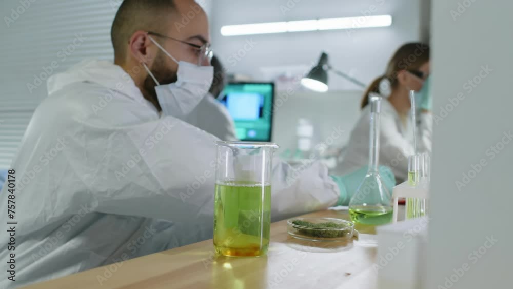 Waist up of focused Middle Eastern male scientist adding red liquid into yellow mixture in glass beaker with pipette while doing research at table in modern lab Waist up of focused Middle Eastern male scientist adding red liquid into yellow mixture in glass beaker with pipette while doing research at table in modern lab
