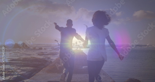 Bright spot of light against rear view of african american couple running on the rocks near the sea