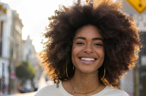 A woman with curly hair is smiling and wearing a gold necklace. She is standing on a street with a car behind her