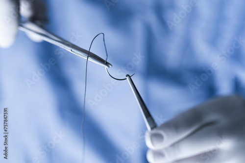 Closeup photo of a medical professional in blue scrubs handling sutures. Operator holding half circle needle with forceps and needle holder during operation.