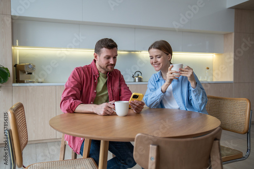 Smiling millennial couple at table kitchen have fun using smartphone together. Relaxed lovers man and woman enthusiastically watch funny video on cellphone screen over coffee spend good time at home.