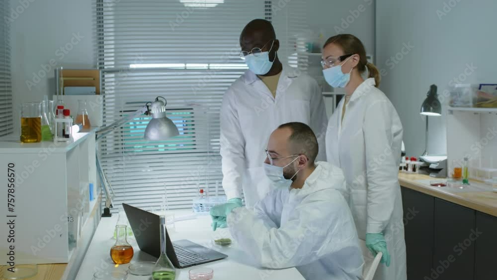 Medium shot of multiracial team of scientists in lab coats and face masks looking at laptop screen and having discussion while working on research together in modern laboratory Medium shot of multiracial team of scientists in lab coats and face masks looking at laptop screen and having discussion while working on research together in modern laboratory