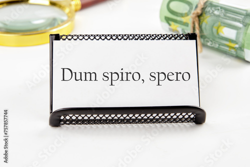 Dum Spiro Spero - latin phrase means While I Breath, I Hope. on a white business card on a white background in a black stand next to it in non-focus are money and a magnifying glass photo