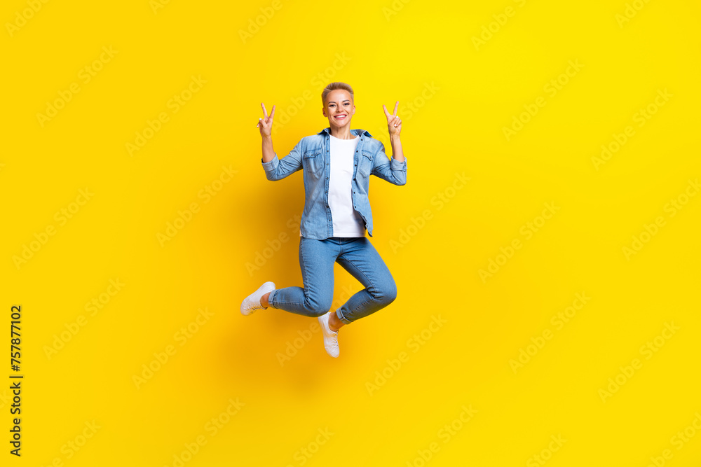 Full size photo of pretty young girl jumping show v-sign hello wear trendy jeans outfit isolated on yellow color background