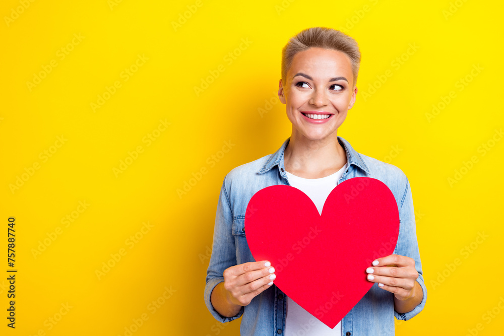 Photo portrait of pretty young girl hold big red heart look empty space wear trendy jeans outfit isolated on yellow color background Photo portrait of pretty young girl hold big red heart look empty space wear trendy jeans outfit isolated on yellow color background