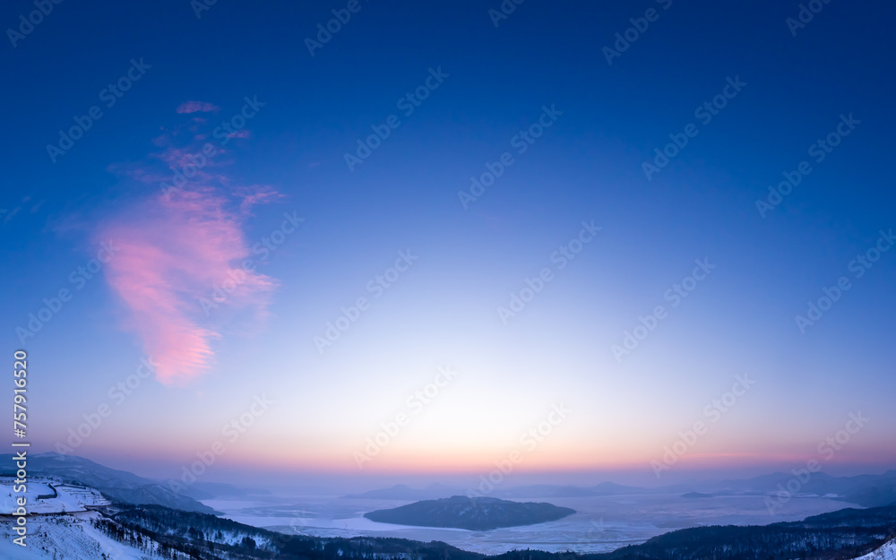 Obraz premium 広い夜明けの空の下の朝もやに霞んだ峰から見下ろす結氷した湖。北海道の美幌峠から見る屈斜路湖。