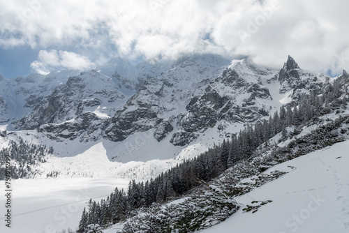 Winter Tatra mountains panorama with snowy trees and frozen Morkie Oko. High rocky mountains covered with clouds lighteen by sun and Mnich (Monk) peak in the background. © Marcin Mucharski