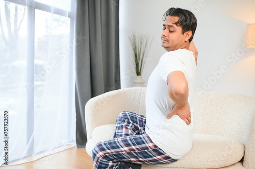 A sick man on Christmas has severe back pain, massages muscle his hand sitting on sofa in living room