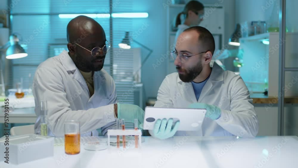 Waist up of two ethnically diverse male lab scientists looking at research observations on tablet computer and having discussion while conducting experiment at table in modern laboratory Waist up of two ethnically diverse male lab scientists looking at research observations on tablet computer and having discussion while conducting experiment at table in modern laboratory