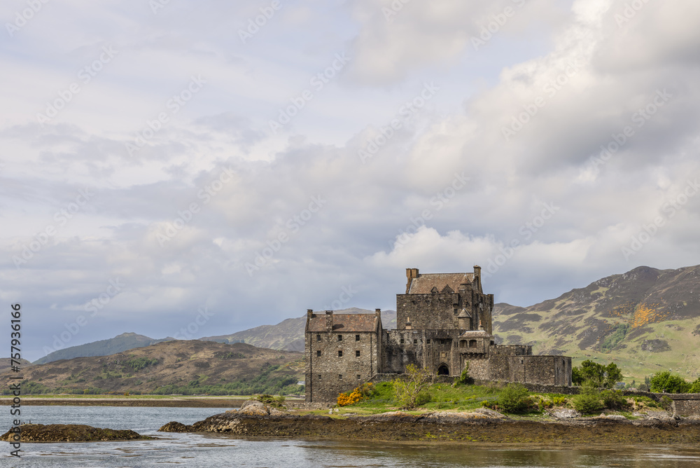 Clouds drift above Eilean Donan Castle, a monument of Scottish heritage poised on the water's edge, framed by the rolling hills of the Highlands