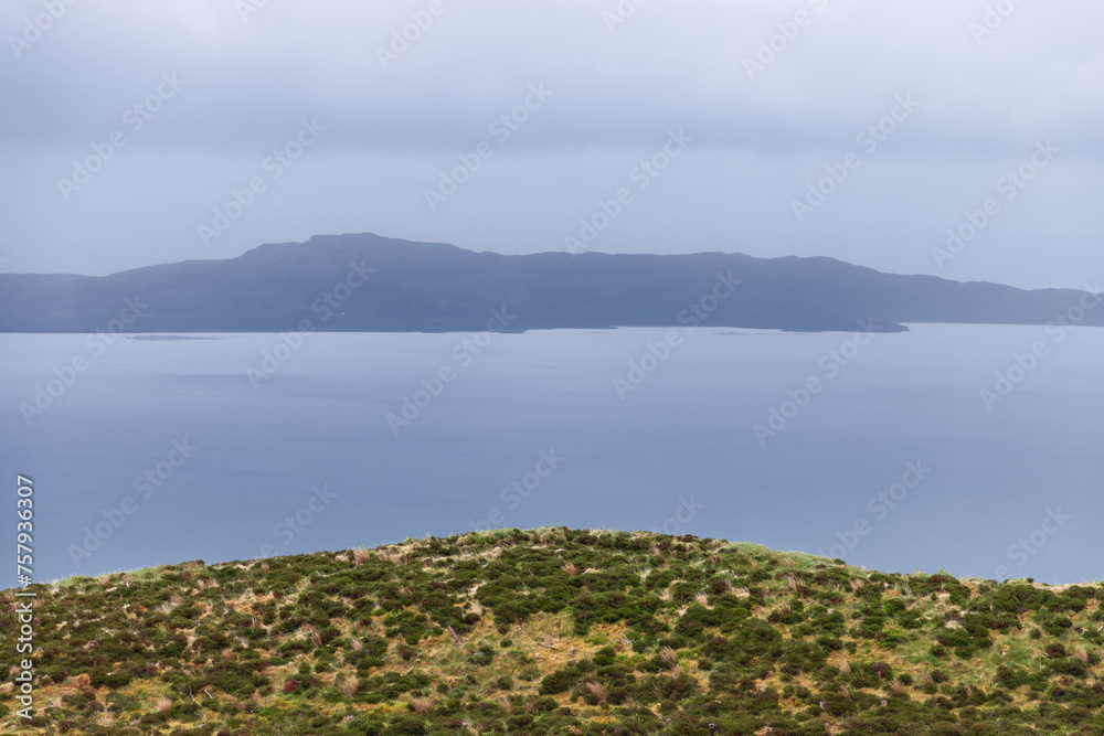 A serene expanse of sea blurs with the hazy outline of Raasay Island under a soft grey sky, framed by the wild heath of the Isle of Skye