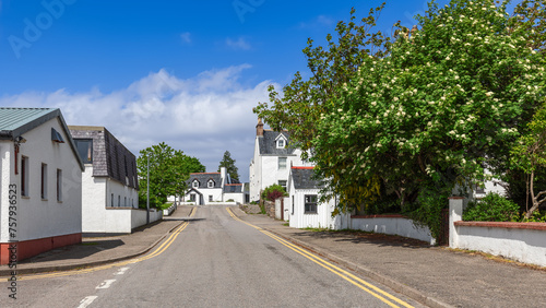 The quaintness of Ullapool is on display with white-rendered houses nestled among vibrant green foliage and flowering trees, beneath a blue sky. Highland Council, Scotland