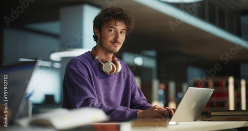 Portrait of a Handsome European Student Studying in a Traditional Library. Young Male Looking at Camera and Smiling. Scholar Working on Laptop Computer in College, Researching Academic Topics Online