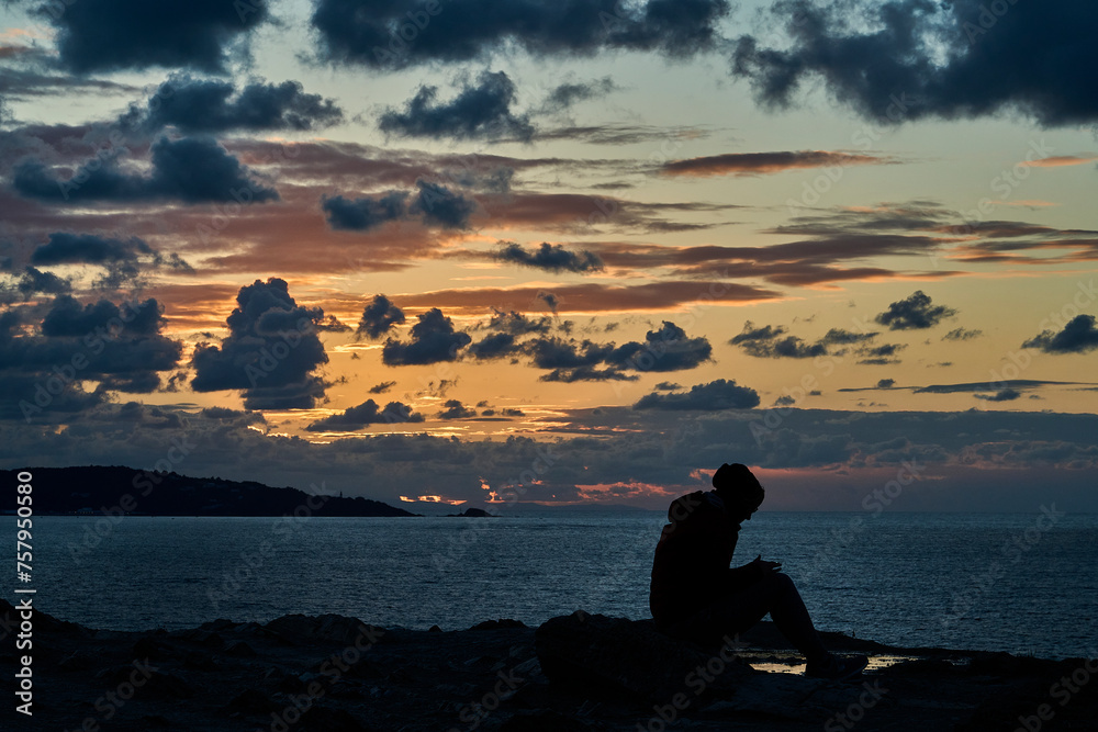 unrecognizable girl in silhouette sitting at the edge of steep and rugged cliffs of the gulf of biscay bay near St. Jean de Luz and using her smart phone with dramatic sunset and cloudscape.