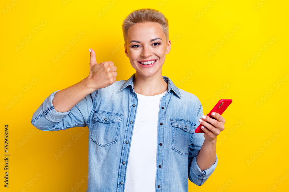 Photo portrait of attractive young woman hold gadget show thumb up dressed stylish denim clothes isolated on yellow color background