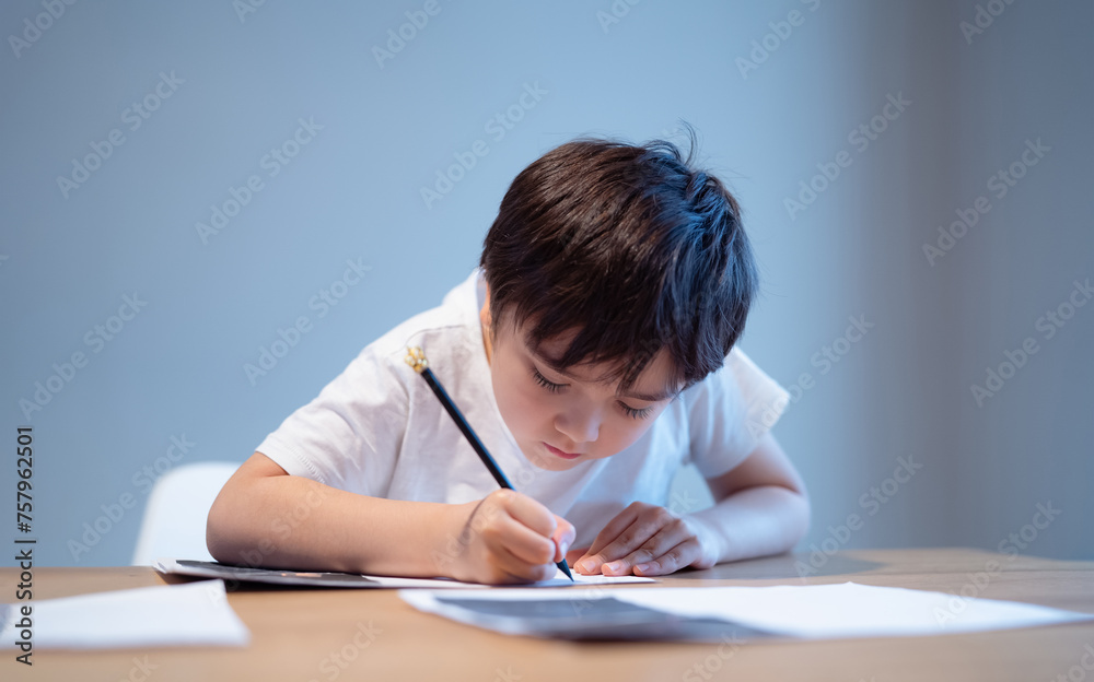 Kid siting on table doing homework,Child boy holding black pen writing on white paper,Young boy practicing English words at home. Elementary school and home schooling, Distance Education concept Kid siting on table doing homework,Child boy holding black pen writing on white paper,Young boy practicing English words at home. Elementary school and home schooling, Distance Education concept