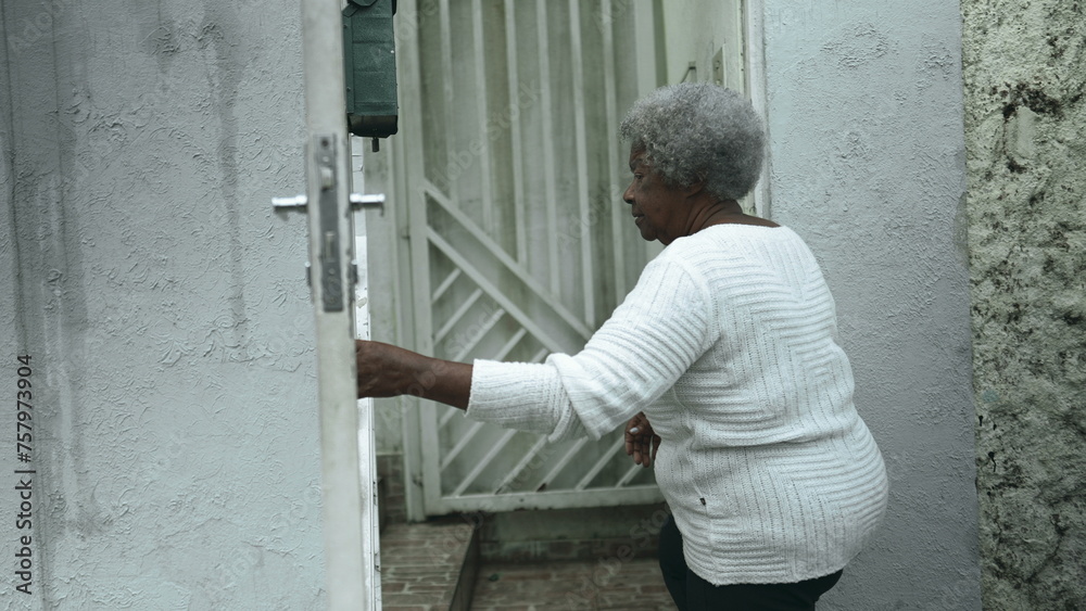 Elderly South American black woman arrives home from sidewalk street, opens residence front door returning by closing gate behind her, senior 80s person of African descent