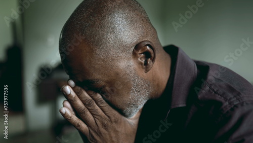 One depressed senior man covers face in shame and regret in moody gloomy bedroom. African American person struggles with poverty and mental illness, close-up face feeling hopeless