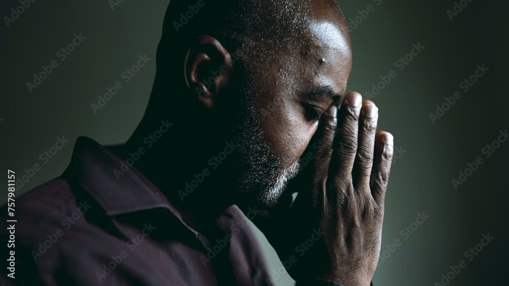 One religious black senior man in contemplative PRAYER at home in dimly lit room with eyes closed having HOPE and FAITH during challenging times. Spiritual African American 50s person - obrazy, fototapety, plakaty 