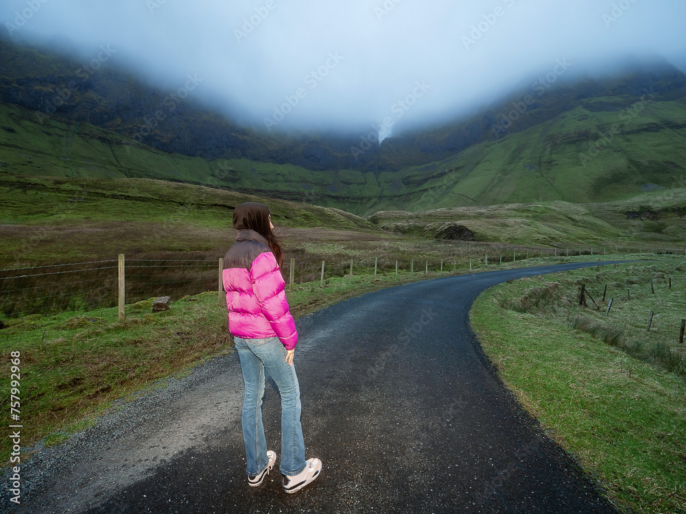 Teenager girl standing on a small country road with stunning nature scenery around her. Tall mountains with grass fields under low cloudy sky. Horseshoe loop drive, county Sligo, Ireland Travel theme