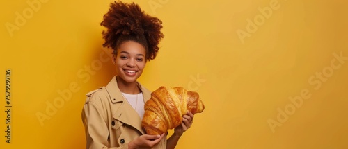 African young woman with big bun haircut wearing trench holding huge croissant, isolated orange background