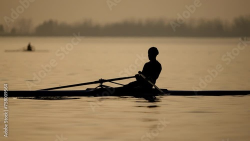 The silhouette of a rowing athlete in a boat training on the lake against the background of golden reflections of the water at sunset