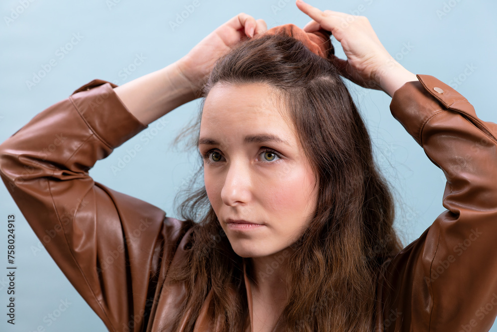 Naklejka premium Portrait of the female person in a brown eco-leather dress making hairstyle herself. A woman is preparing to go to a meeting.