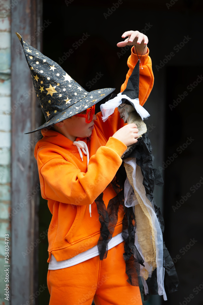Fototapeta premium A child in an orange costume and a wizard hat for Halloween. A boy holds a skeleton at a Halloween party.