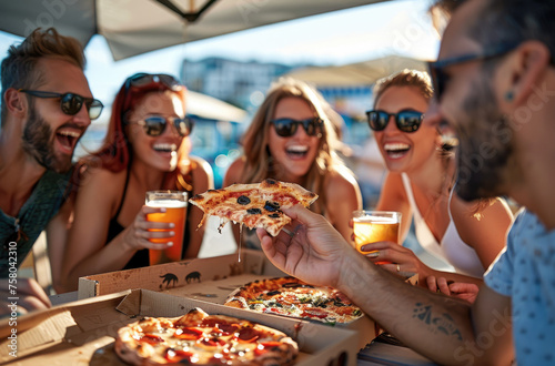Happy friends in sunglasses eating pizza and drinking beer at a rooftop party, laughing while sitting at a table with a box of pizzeria food