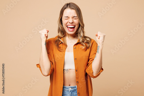 Young Caucasian woman she wears orange shirt casual clothes doing winner gesture celebrate clenching fists say yes close eyes isolated on plain pastel light beige background studio. Lifestyle concept.