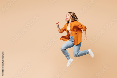 Full body side profile view excited happy young woman wear orange shirt casual clothes jump high run fast hurry up isolated on plain pastel light beige background studio portrait. Lifestyle concept.